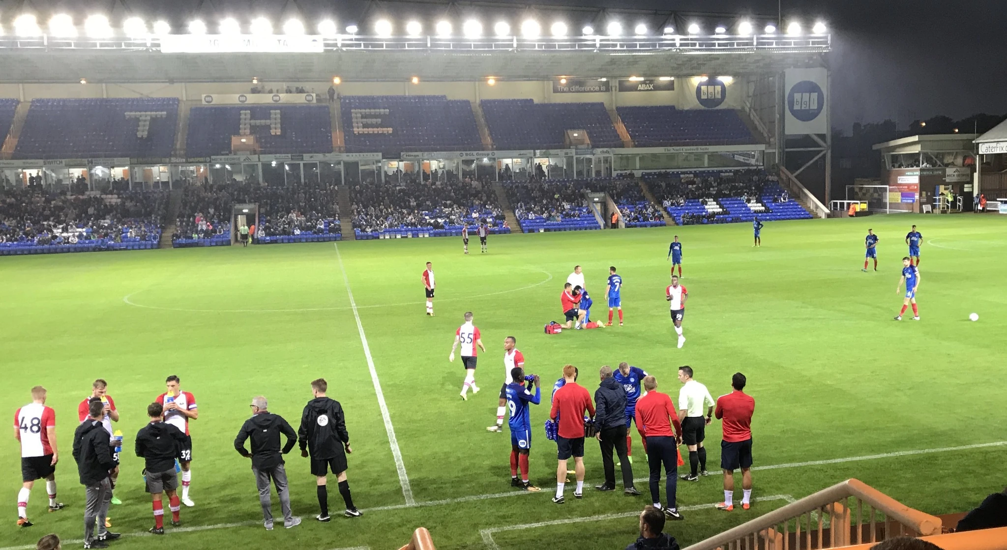 A stop in play as a Peterborough United player receives attention on the pitch, whilst Martin Hunter and Kelvin Davis offer advice to the Southampton players on the left of the picture, and Peterborough United Manager Grant McCann and Assistant David Oldfield talk with their players by the Technical area.